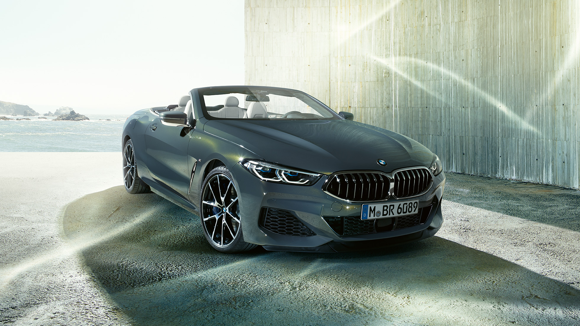 BMW M850i xDrive Cabrio, trīs ceturtdaļu skats no priekšpuses. BMW M850i xDrive Competition Cabrio metāliskā Dravit Grey krāsā ar nolaistu mīkstā seguma jumtu, trīs ceturtdaļu skats no sāniem, stāvošs automobilis uz piekrastes fona.