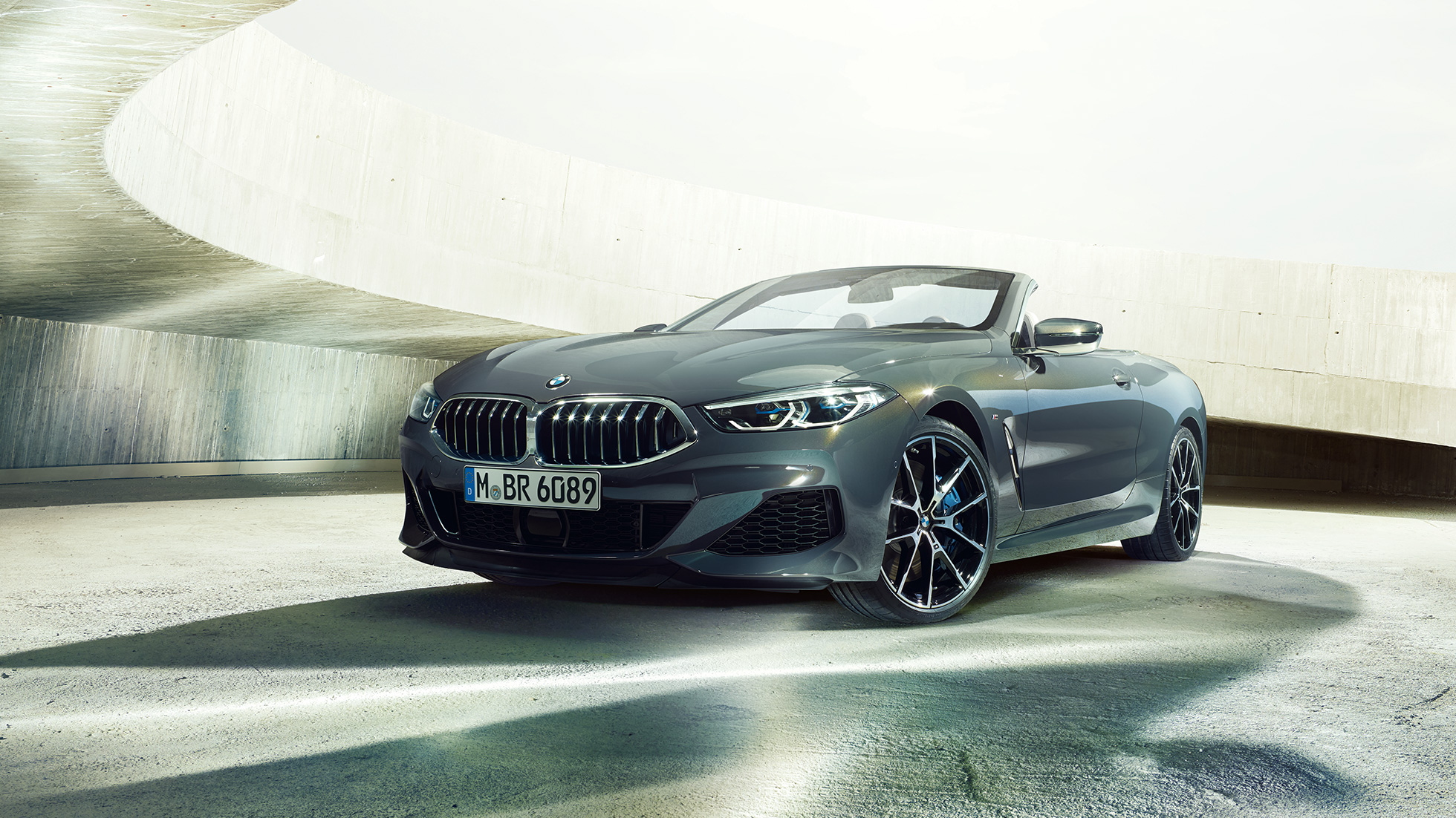 BMW M850i xDrive Cabrio, trīs ceturtdaļu skats no priekšpuses. BMW M850i xDrive Competition Cabrio metāliskā Dravit Grey krāsā ar nolaistu mīkstā seguma jumtu, trīs ceturtdaļu skats no priekšpuses, stāvošs automobilis betona ēkā.