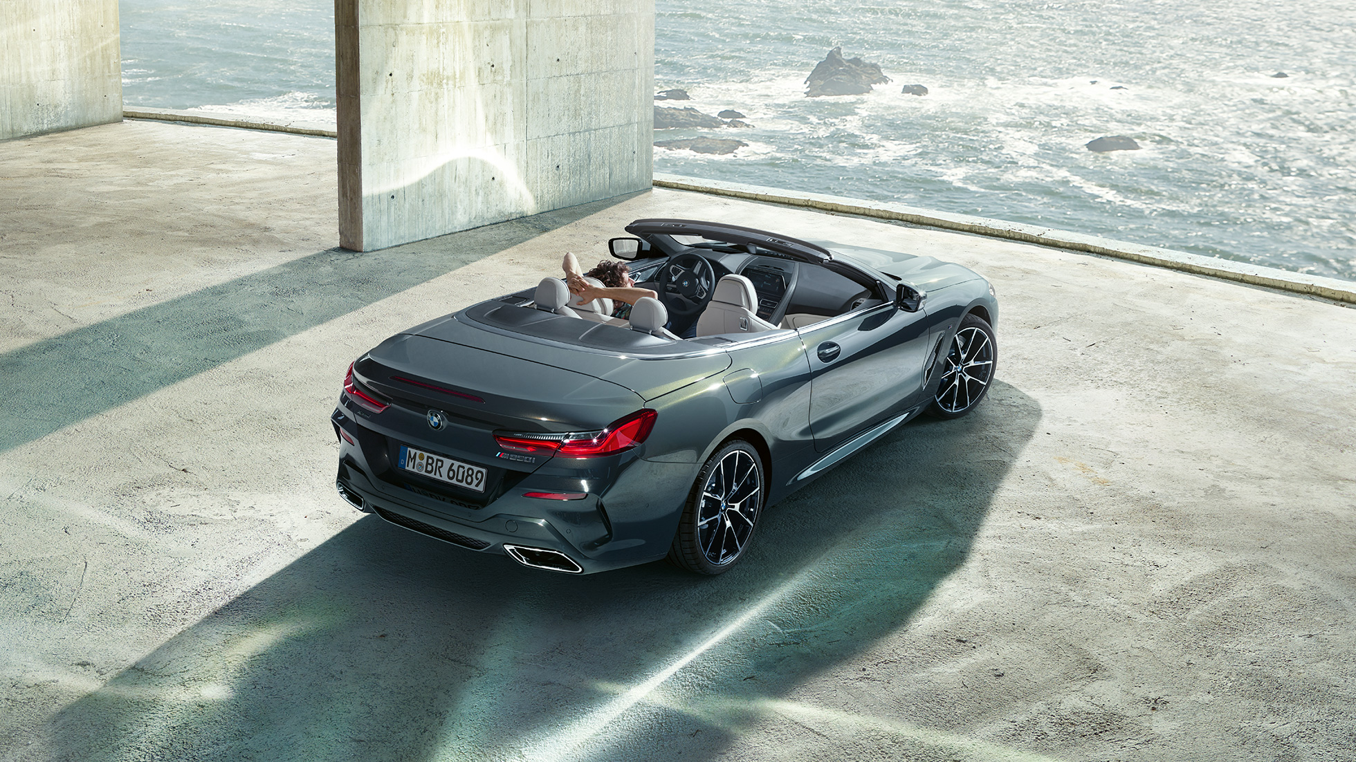 BMW M850i M Cabrio, skats no putna lidojuma. BMW M850i xDrive Cabrio metāliskā Dravit Grey krāsā ar nolaistu mīkstā seguma jumtu, skats no putna lidojuma, stāvēšanai novietots automobilis betona ēkā uz okeāna fona.