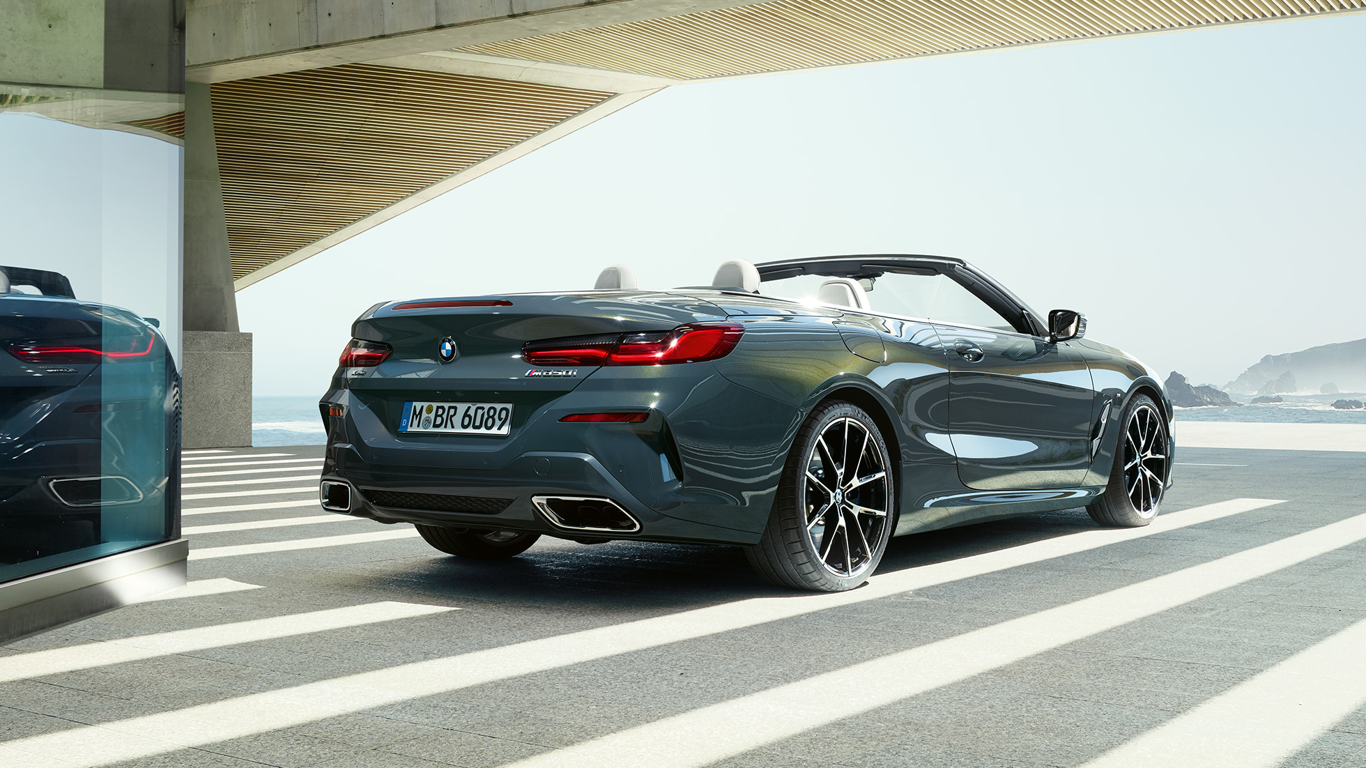 BMW M850i xDrive Cabrio, trīs ceturtdaļu skats no aizmugures. BMW M850i xDrive Competition Cabrio metāliskā Dravit Grey krāsā ar nolaistu mīkstā seguma jumtu, trīs ceturtdaļu skats no aizmugures uz novietotu automobili uz okeāna ainava fona.