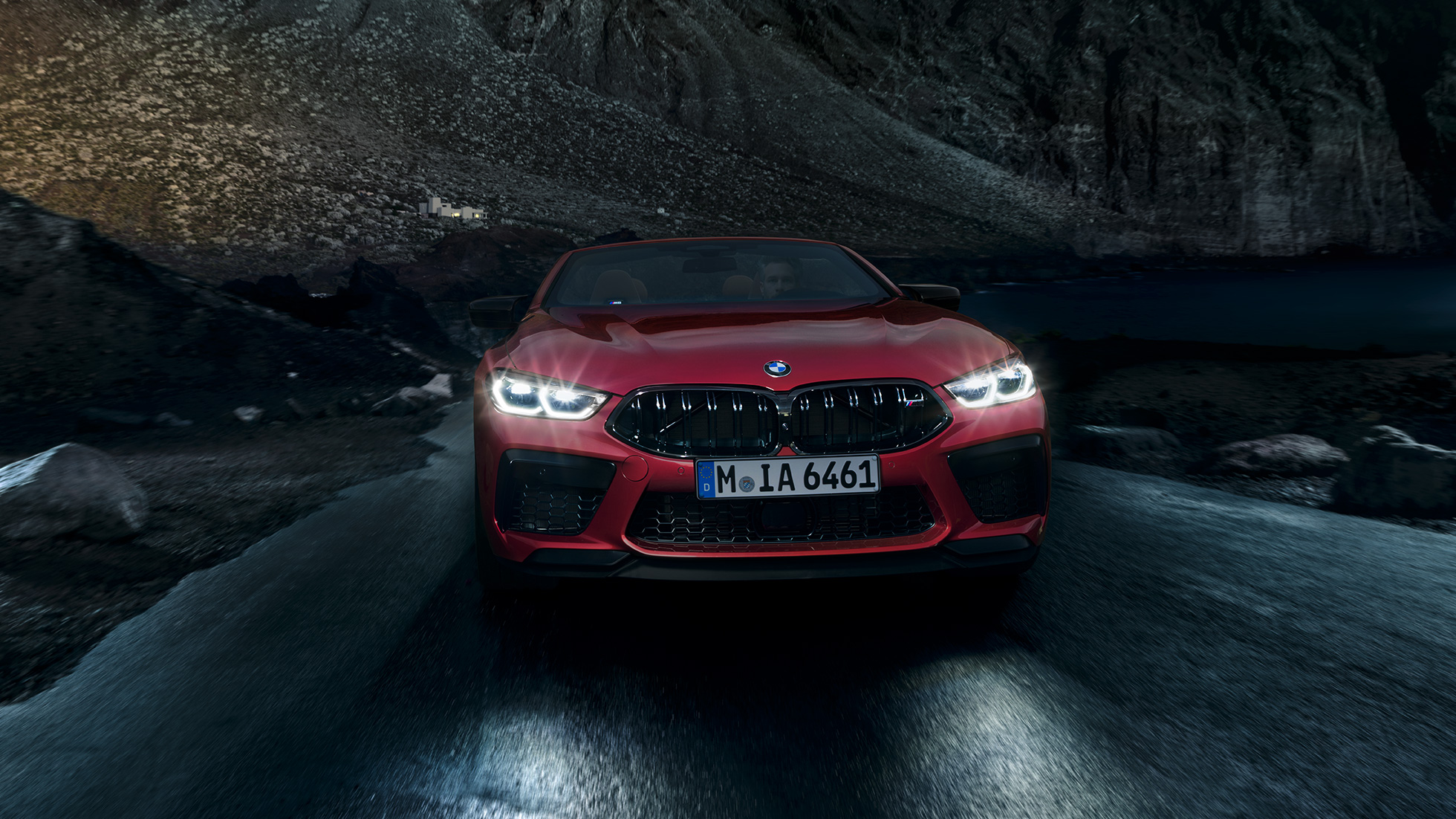 BMW M8 Competition Cabrio, skats no priekšpuses. BMW M8 Competition Cabrio metāliskā Motegi Red krāsā ar ieslēgtiem priekšējiem lukturiem, skats no priekšpuses, braucošs automobilis naktī.