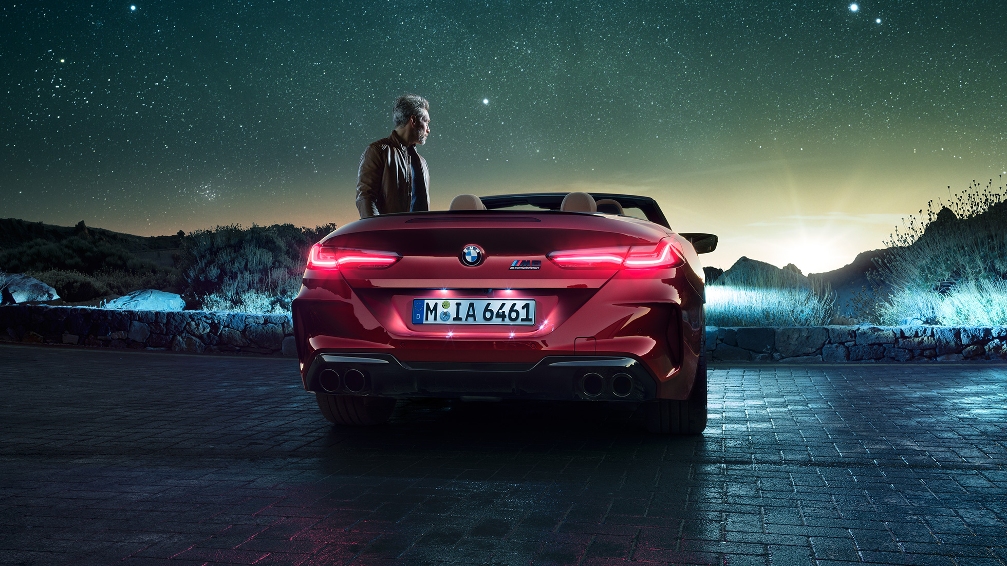BMW M8 Competition Cabrio, skats no aizmugures. BMW M8 Competition Cabrio metāliskā Motegi Red krāsā ar nolaistu mīkstā seguma jumtu, skats no aizmugures, stāvošs automobilis ar vīrieti zem zvaigžņotām debesīm.