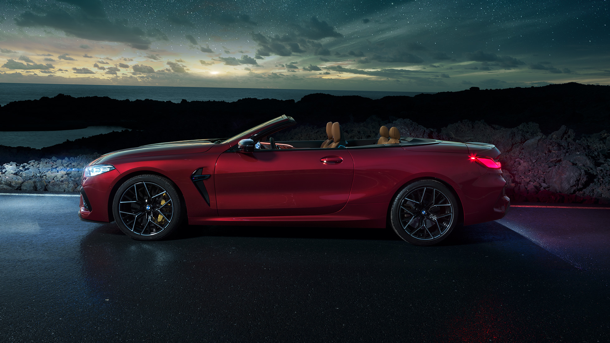 BMW M8 Competition Cabrio, skats no sāniem. BMW M8 Competition Cabrio metāliskā Motegi Red krāsā ar nolaistu mīkstā seguma jumtu, skats no sāniem, stāvošs automobilis ar zem zvaigžņotām debesīm.