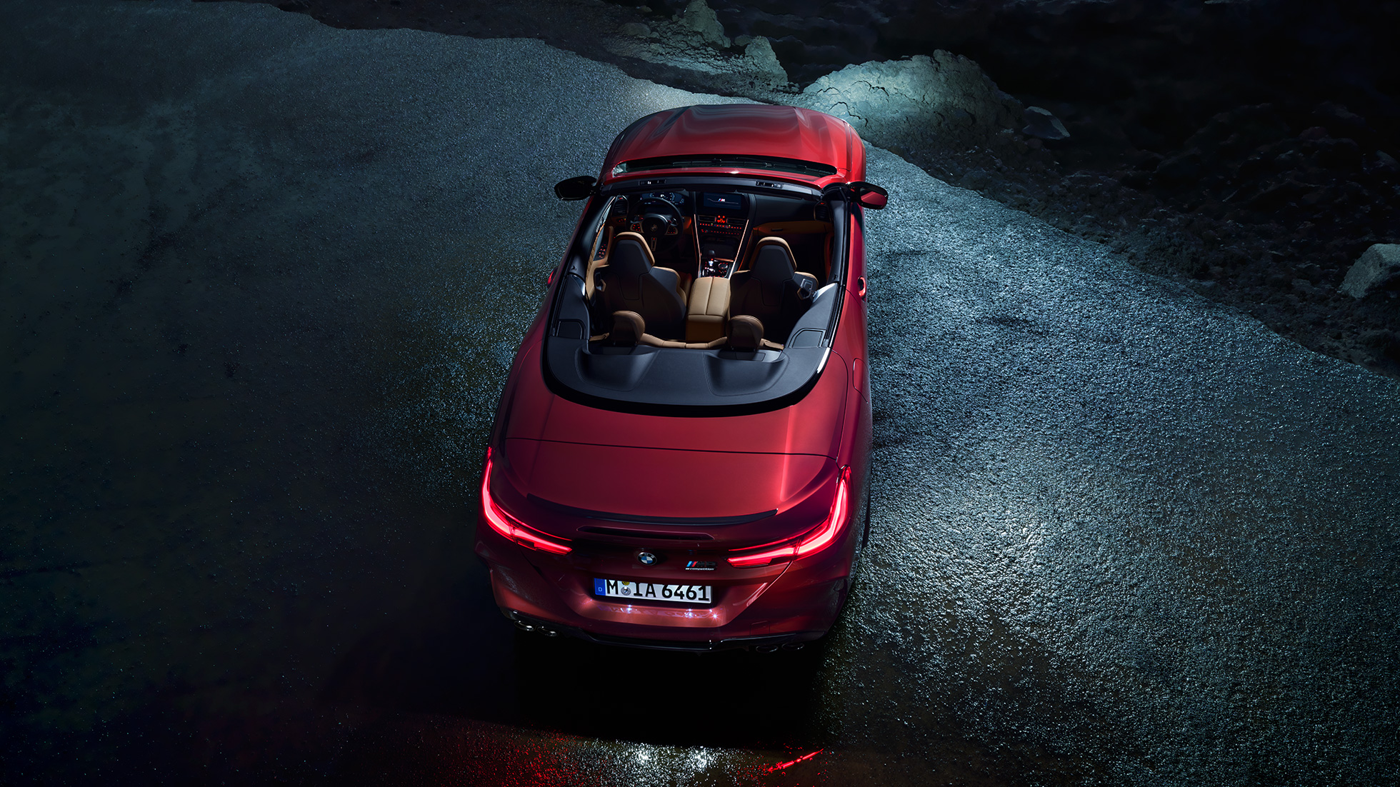 BMW M8 M Competition Cabrio, skats no putna lidojuma. BMW M8 Competition Cabrio metāliskā Motegi Red krāsā ar nolaistu mīkstā seguma jumtu, skats no putna lidojuma, stāvošs automobilis uz asfalta seguma naktī.