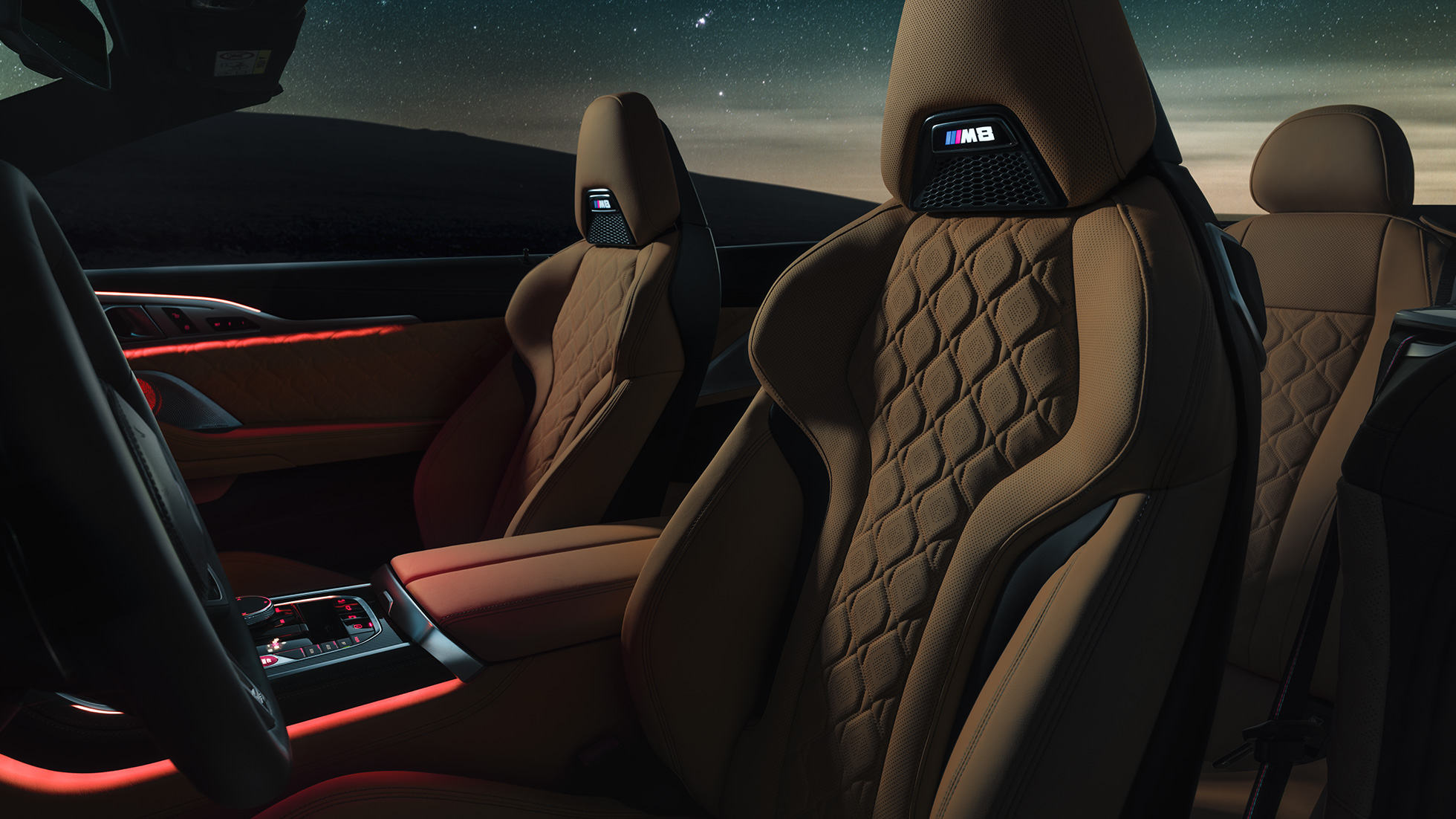 Merino ādas M sporta sēdekļi. BMW M8 Competition Cabrio ar nolaistu mīkstā seguma jumtu zem zvaigžņotām debesīm, salons ar M sporta sēdekļiem ar Merino ādu un sarkanu fona apgaismojumu.