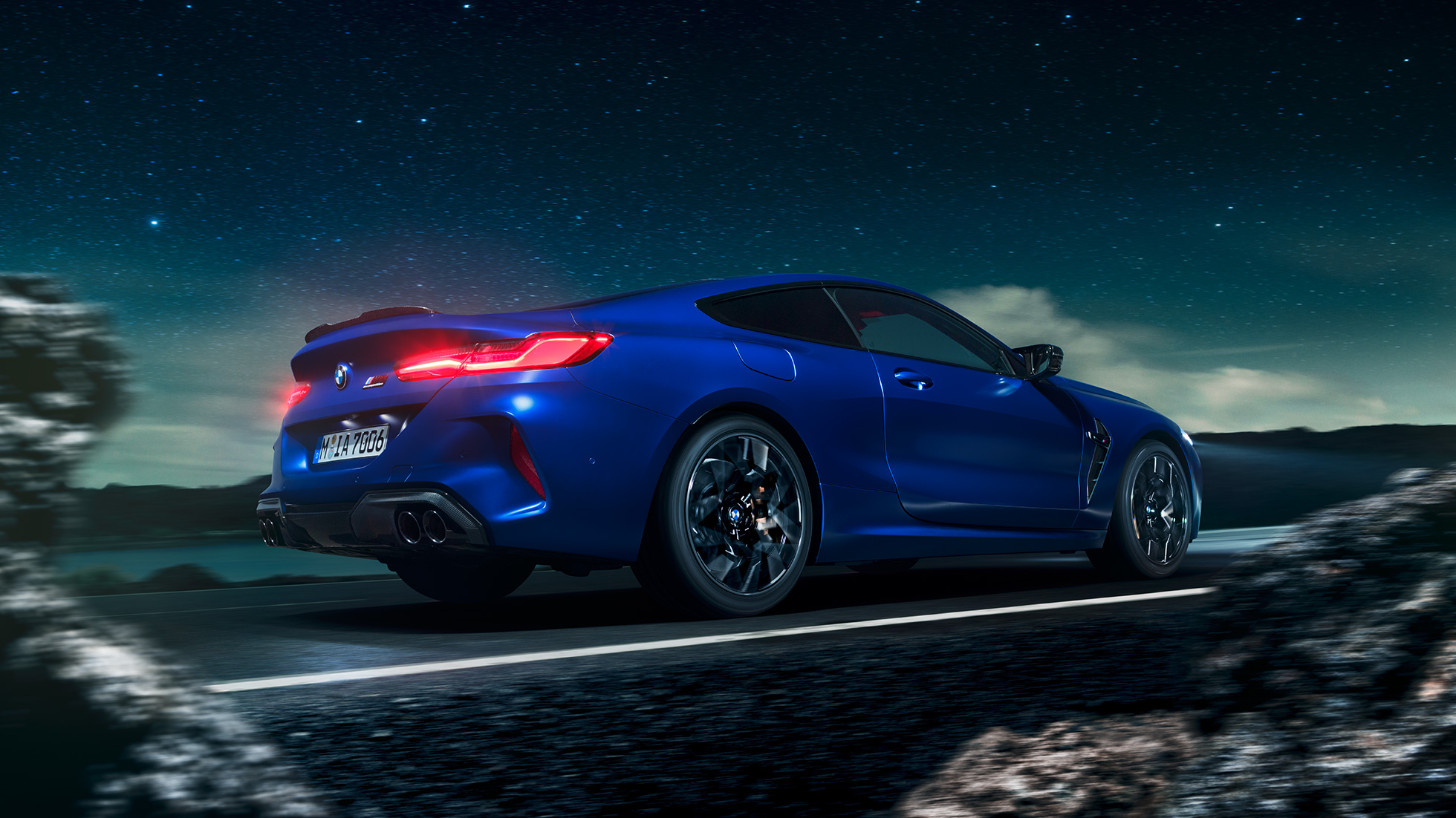 BMW M8 M Competition Coupé, trīs ceturtdaļu skats no sāniem. BMW M8 Competition Coupé metāliskā Marina Bay Blue krāsā, trīs ceturtdaļu skatā no priekšpuses uz braucošu automobili uz zvaigžņotas debess fona.