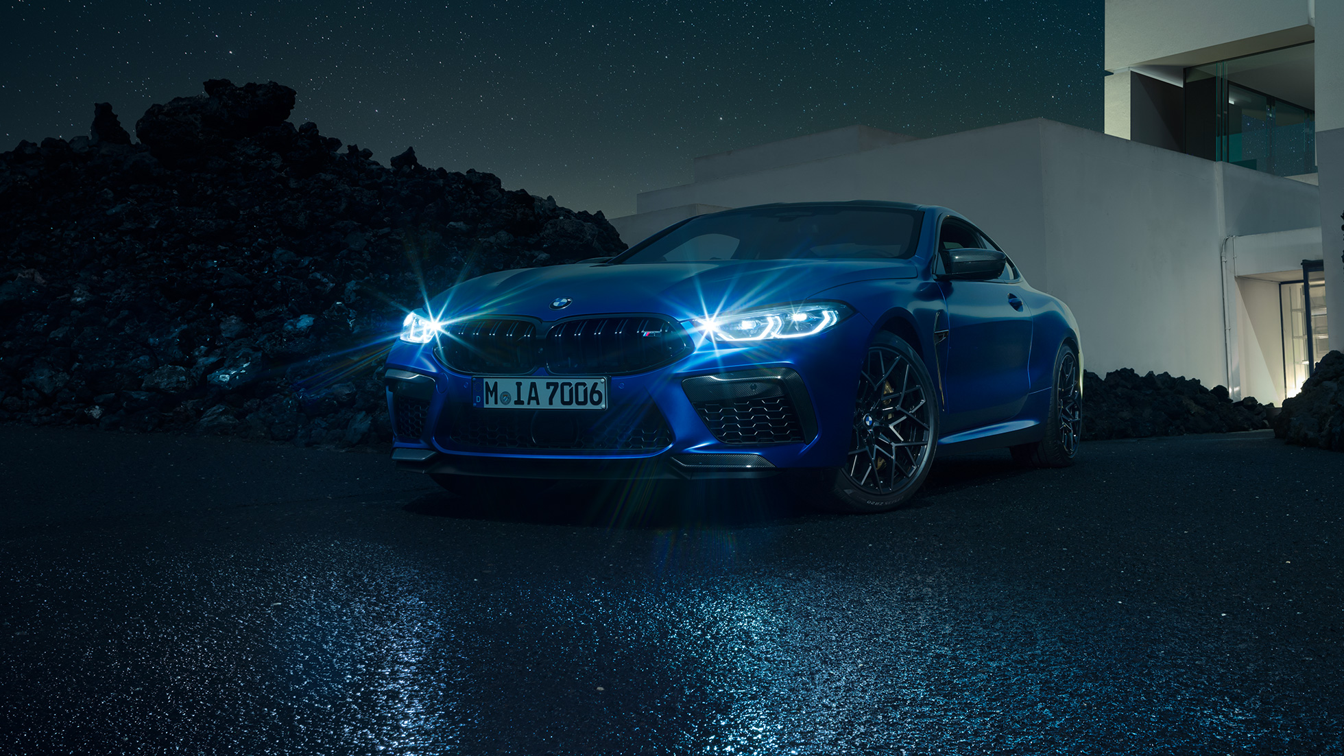BMW M8 M Competition Coupé, trīs ceturtdaļu skats no priekšpuses. BMW M8 Competition Coupé metāliskā Marina Bay Blue krāsā ar ieslēgtiem LED priekšējiem lukturiem, trīs ceturtdaļu skatā no priekšpuses uz villas priekšā novietotu automobili naktī.