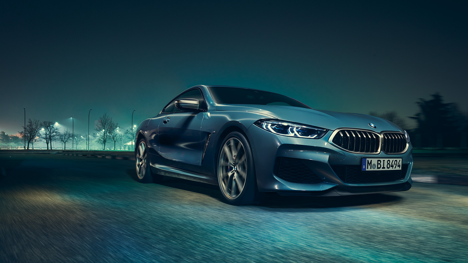 BMW M850i xDrive Coupé, priekšpuse trīs ceturtdaļu skatā no kreisās puses. BMW M850i xDrive Coupé Coupé metāliskā Barcelona Blue krāsā, priekšpuse trīs ceturtdaļu skatā no kreisās puses ar braucošu automobili naktī.