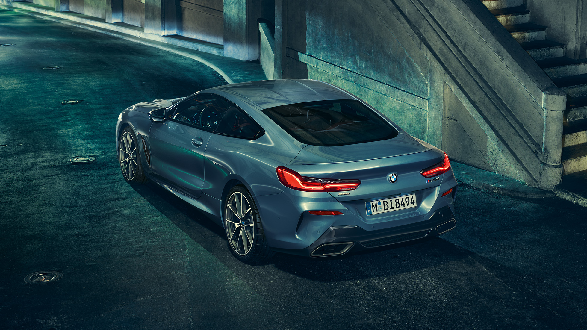 BMW M850i xDrive Coupé, aizmugure trīs ceturtdaļu skatā no kreisās puses. BMW M850i xDrive Coupé Coupé metāliskā Barcelona Blue krāsā, priekšpuse trīs ceturtdaļu skatā no kreisās puses ar stāvošu automobili naktī.