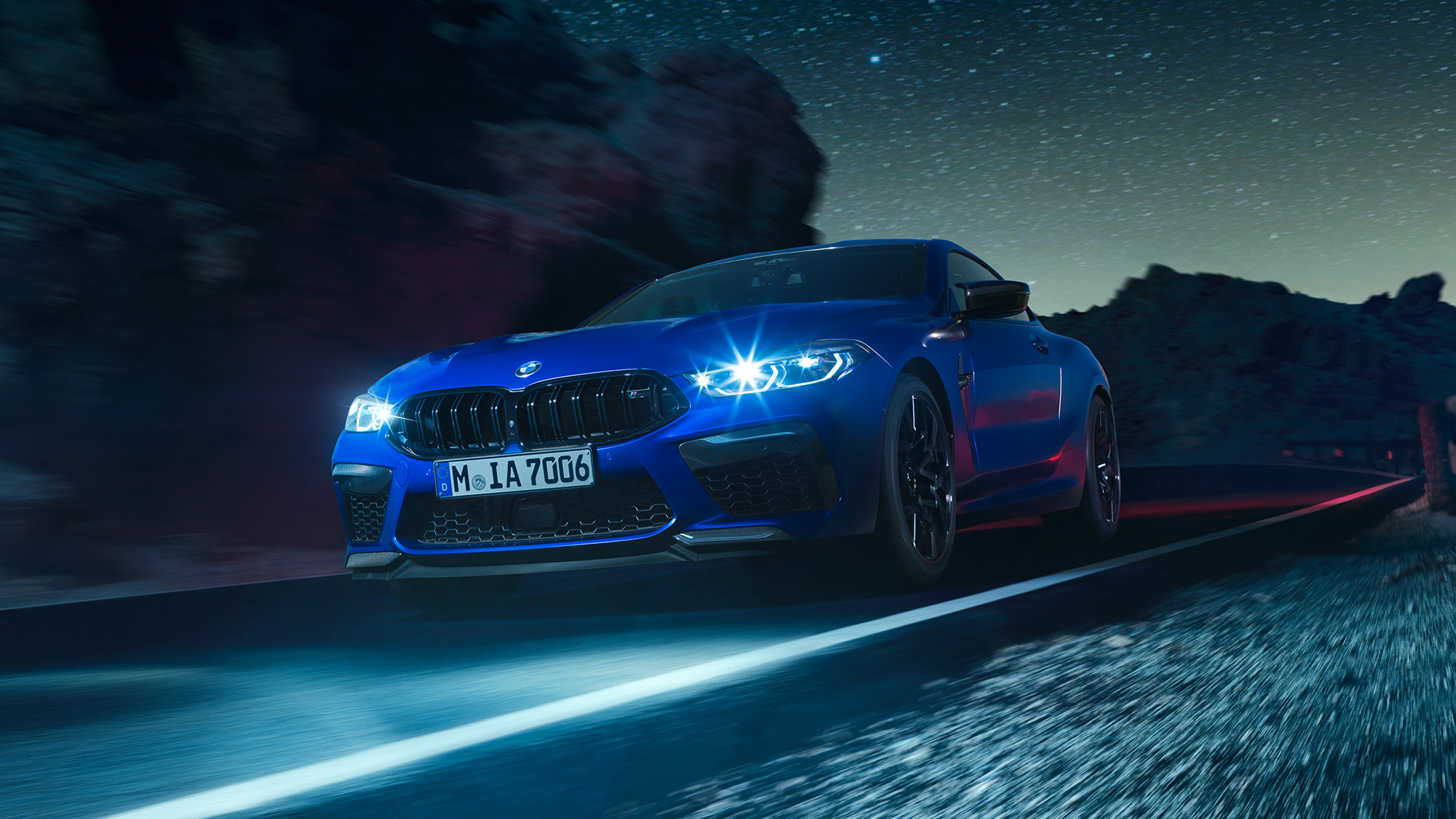 Braucošs BMW M8 M Competition Coupé, trīs ceturtdaļu skats no priekšpuses. BMW M8 Competition Coupé metāliskā Marina Bay Blue krāsā ar iedegtiem LED priekšējiem lukturiem, trīs ceturtdaļu skatā no priekšpuses uz braucošu automobili naktī.