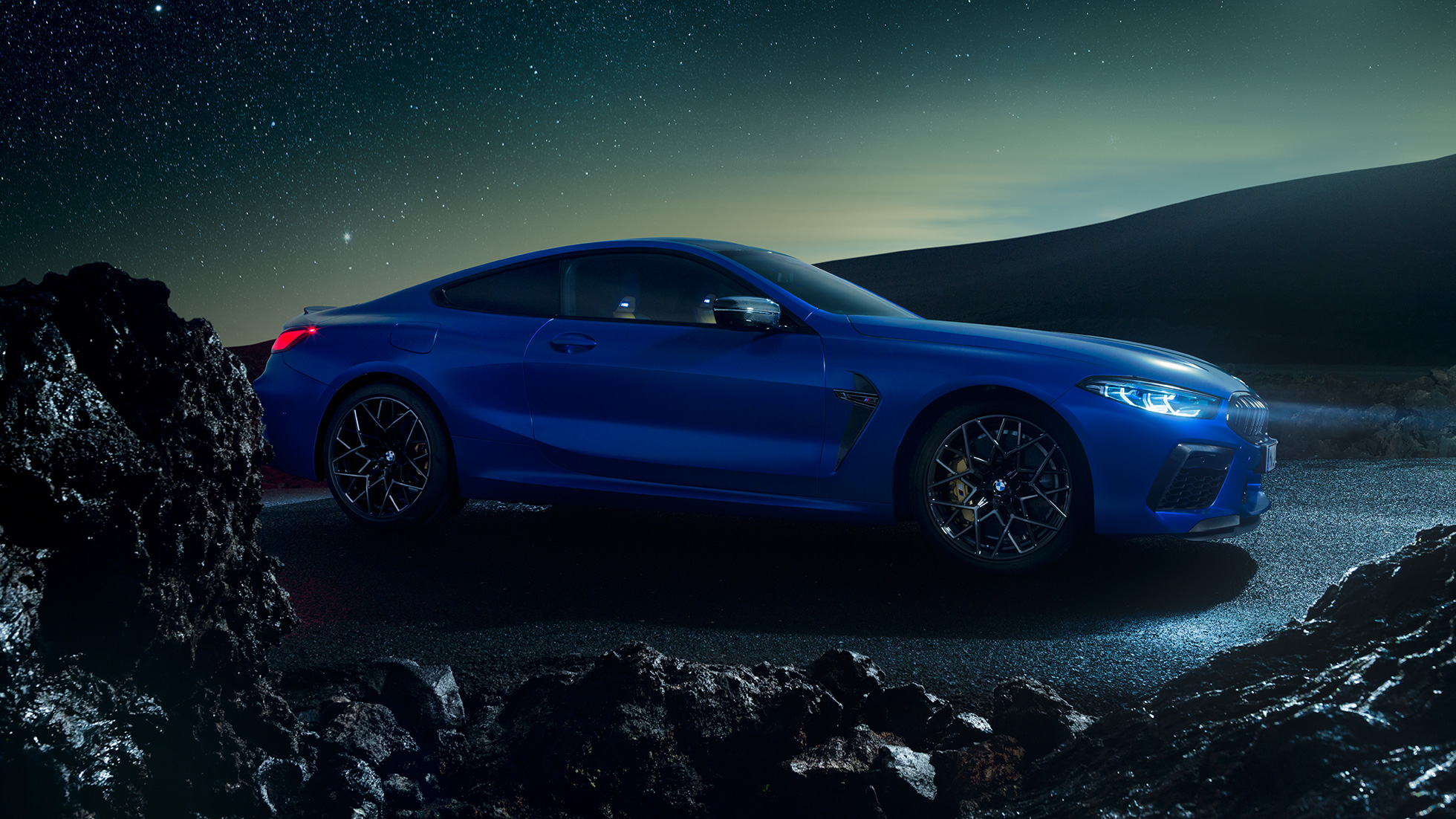 BMW M8 M Competition Coupé, trīs ceturtdaļu skats no sāniem. BMW M8 Competition Coupé metāliskā Marina Bay Blue krāsā, trīs ceturtdaļu skatā no priekšpuses uz stāvošu automobili naktī.