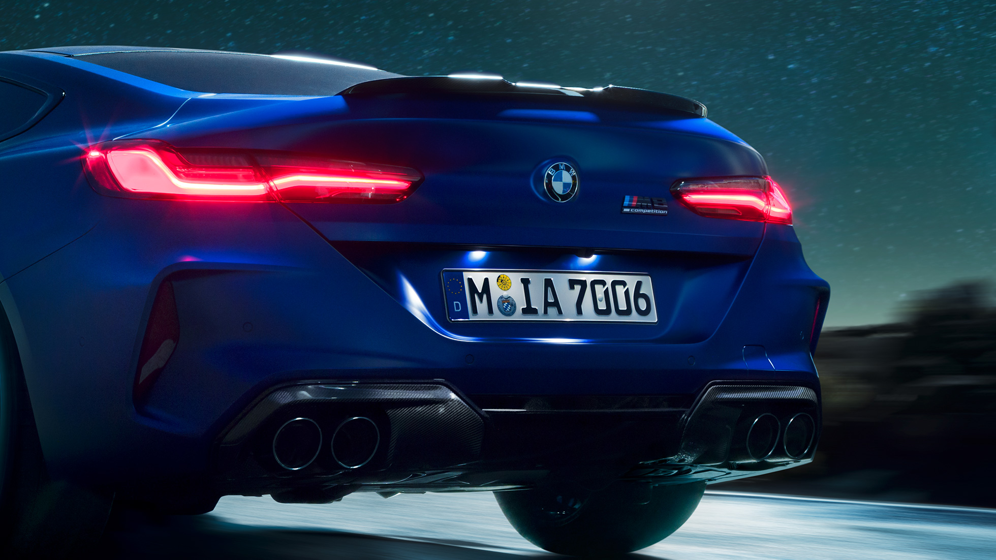 M difuzors Carbon Black krāsā. BMW M8 Competition Coupé metāliskā Marina Bay Blue krāsā, aizmugures tuvplāns naktī ar M difuzoru Carbon Black krāsā.