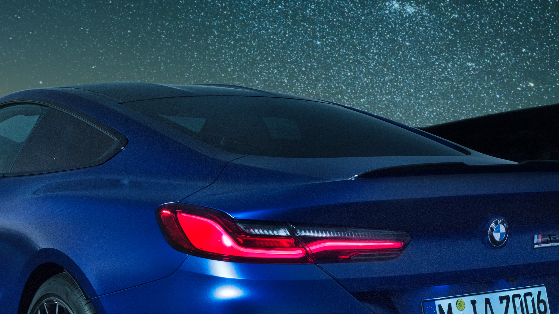 M aizmugurējais spoileris BMW M8 Competition Coupé metāliskā Marina Bay Blue krāsā, naksnīgs plecu daļas tuvplāns ar M aizmugurējo oglekļa spoileri.