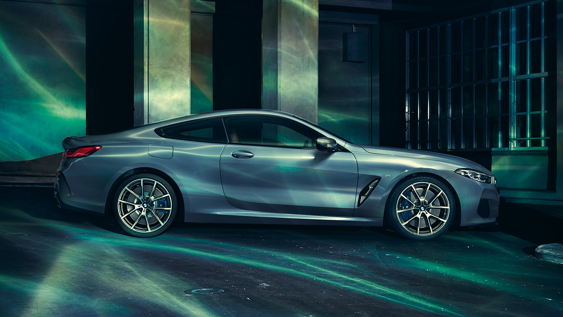 BMW M850i xDrive Coupé, skats no sāniem. BMW M850i xDrive Coupé metāliskā Barcelona Blue krāsā, skats no sāniem uz novietotu automobili uz ēkas fona.