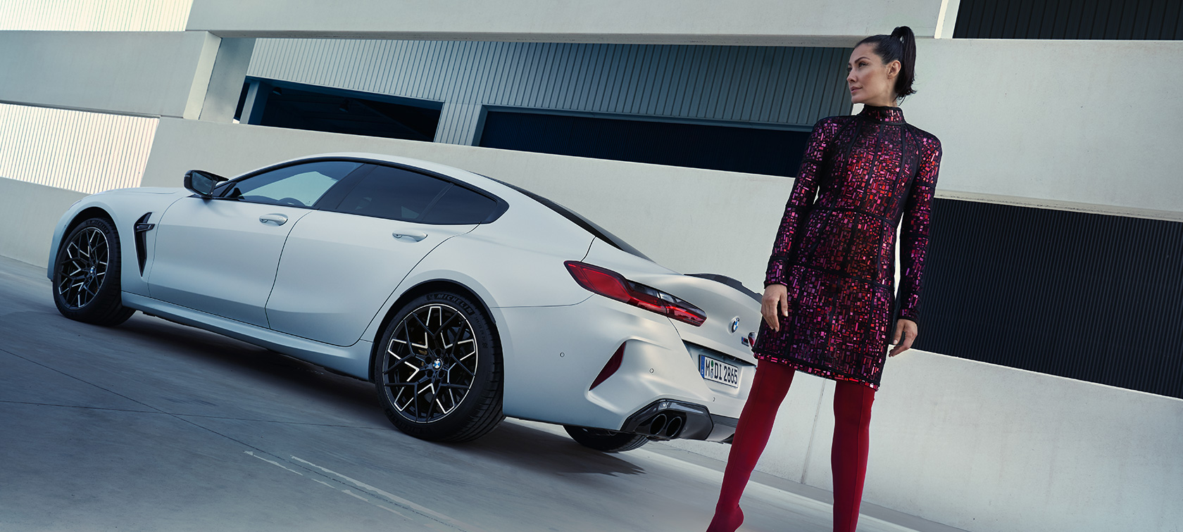 Jaunā BMW M8 Competition Gran Coupé F93, LCI, atjaunināts, 2022, BMW Individual metāliska Brilliant White krāsa, trīs ceturtdaļu aizmugures skats, ar sievieti modeli stāvam blakus automobilim