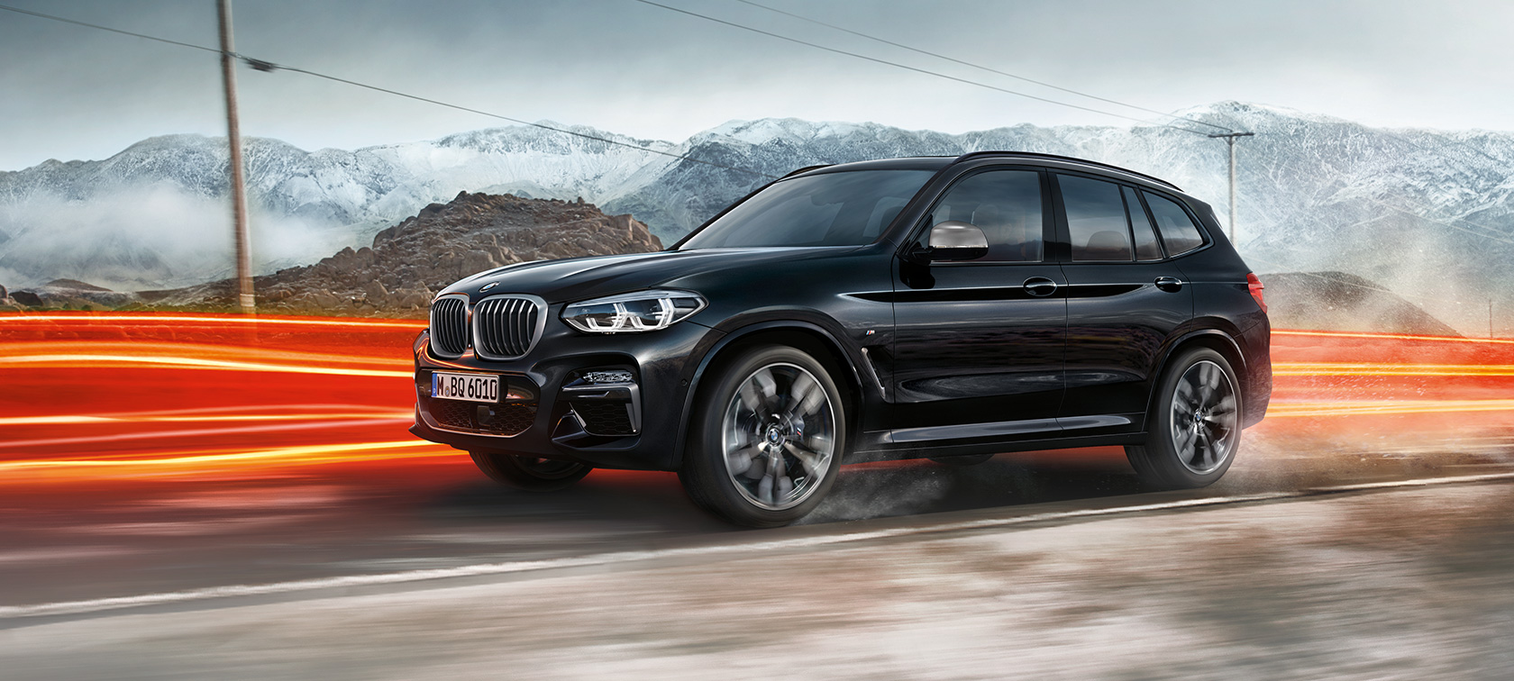 BMW X3 M40i un BMW X3 M40d, skats no sāna. BMW X3 M40i un BMW X3 M40d metāliskā Black Sapphire krāsā, braucošs, virsbūve, skats no sāniem uz kalnu ainavas fona ar gaismas joslām.