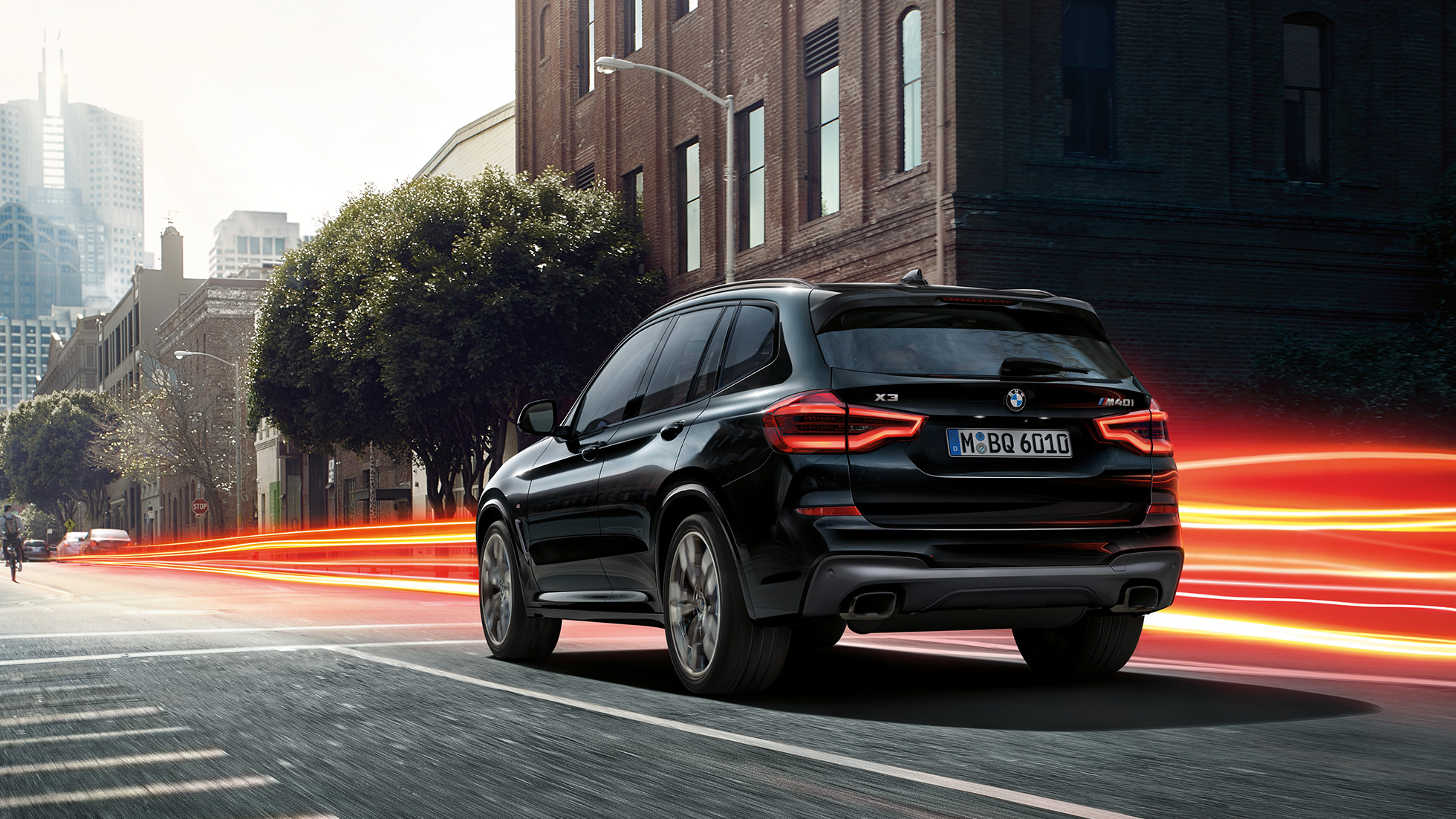 BMW X3 M40i, trīs ceturtdaļu skats no aizmugures. BMW X3 M40i metāliskā Black Sapphire krāsā, braucošs, virsbūve, trīs ceturtdaļu skats no aizmugures uz pilsētas fona ar gaismas joslām.
