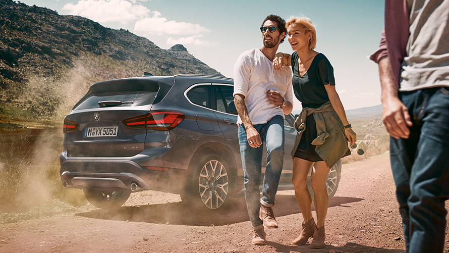 BMW X1, F48, LCI, Storm Bay krāsa, trīs ceturtdaļu aizmugures skats BMW X1, F48, LCI, Storm Bay krāsa, trīs ceturtdaļu aizmugures skats, stāvošs automobilis uz ainavas fona ar cilvēkiem tā priekšā