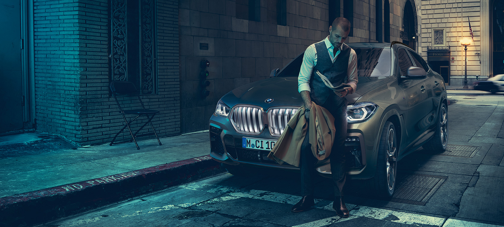 Vīrietis, kas atspiežas pret BMW X6. Vīrietis, kas atspiežas pret BMW X6 skatā no sāna uz pilsētas fona.