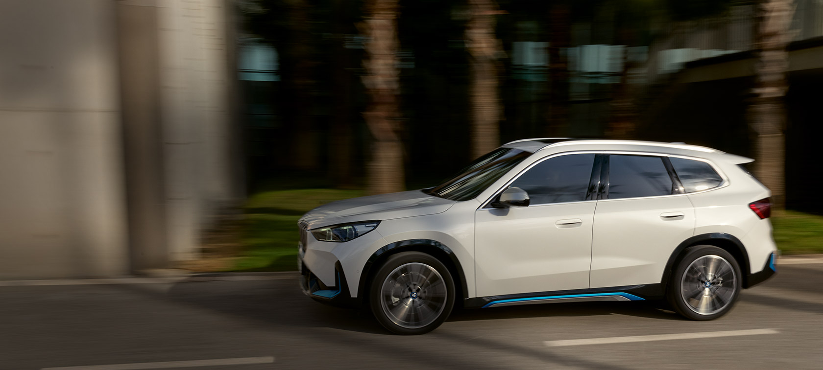 BMW iX1 U11 BEV 2022, metāliska Mineral White krāsa, trīs ceturtdaļu priekšpuses skats, pa kalnu serpentīnu braucošs automobilis