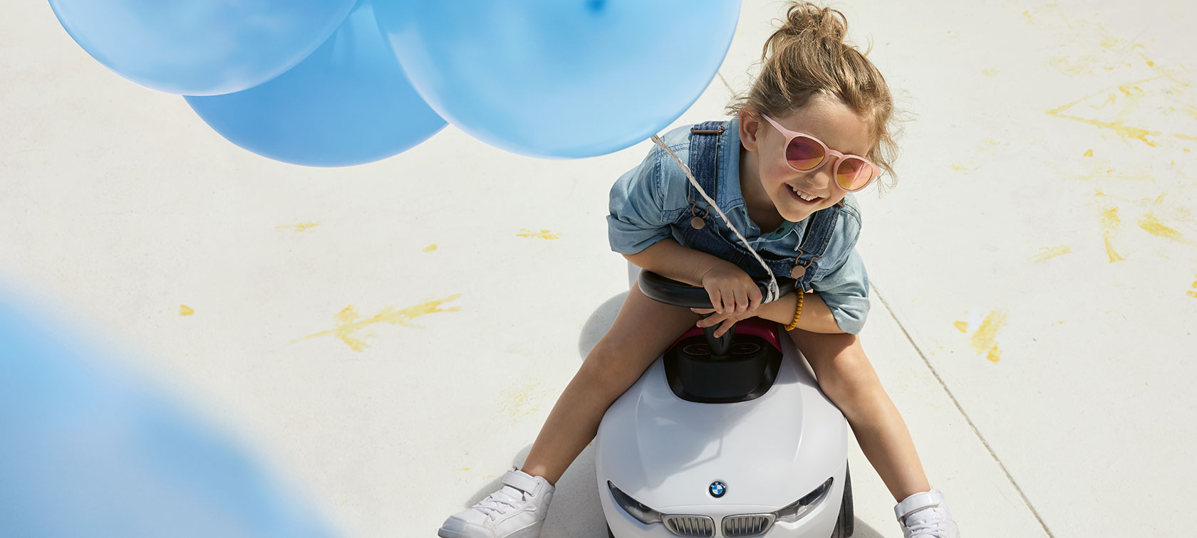 BMW bērniem. Meitene rozā saulesbrillēs un ar zilu balonu priecīgi sēž uz BMW Baby Racer.