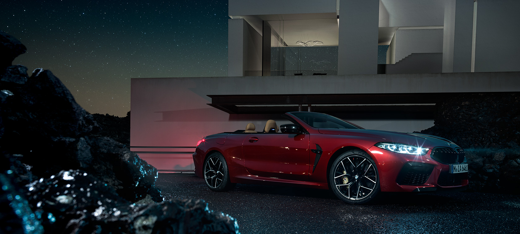 BMW M8 M Competition Cabrio, trīs ceturtdaļu skats no sāniem. BMW M8 Competition Cabrio metāliskā Motegi Red krāsā, trīs ceturtdaļu skats no sāniem, naktī novietots automobilis villas priekšā.
