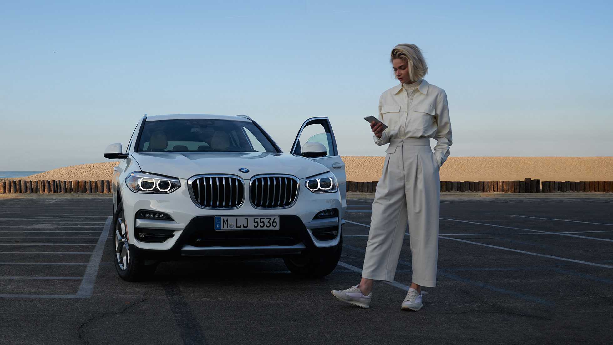 Sieviete darbojas ar My BMW lietojumprogrammu viedtālrunī Sieviete viedtālrunī izmanto BMW ConnectedDrive lietojumprogrammu My BMW uz balta BMW X5 fona