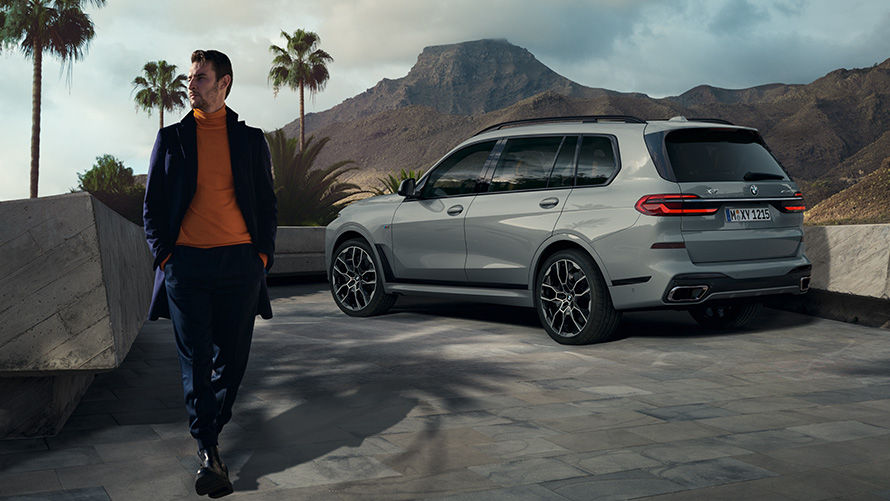 Vīrietis stāvēšanai novietota BMW X7 (G07) priekšā, novietošana stāvēšanai ar tālvadību Novietošana stāvēšanai ar tālvadību, vīrietis stāvēšanai novietota BMW X7 (G07) priekšā, trīs ceturtdaļu aizmugures skats, zem palmu kokiem ar kalnu ainavu fonā