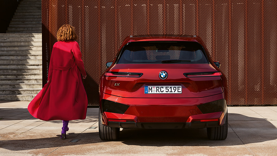 Sieviete sarkanā mētelī blakus BMW iX (i20), bagāžas nodalījuma durvju aizvēršana Bagāžas nodalījuma durvju aizvēršana, sieviete sarkanā mētelī blakus BMW iX (i20), uz ēkas fona