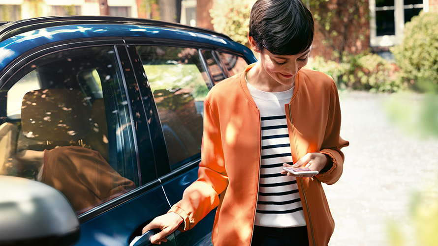 BMW TeleServices Sieviete ar viedtālruni stāv pie sava BMW durvīm.