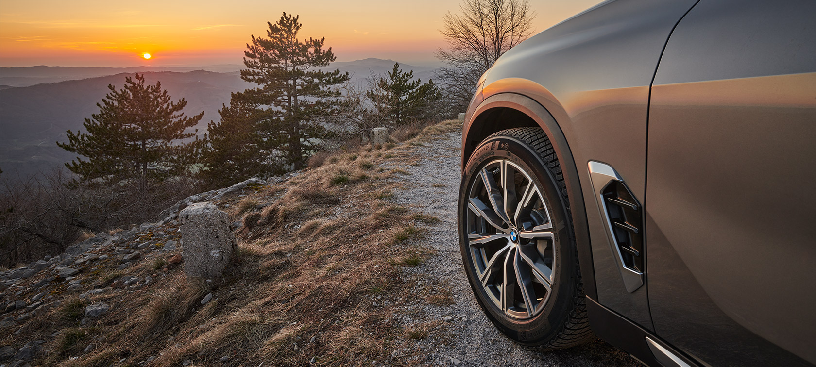 Uz kalna nogāzes fona pa grunts ceļu pretī saulrietam brauc BMW X5 (G05 LCI) ar oriģinālajiem BMW diskiem