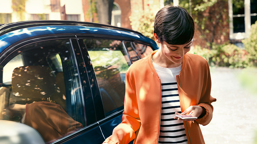 BMW vasaras piedāvājumi, BMW Teleservices BMW vasaras piedāvājumi, BMW Teleservices, sieviete stāv BMW priekšā un izmanto viedtālruni