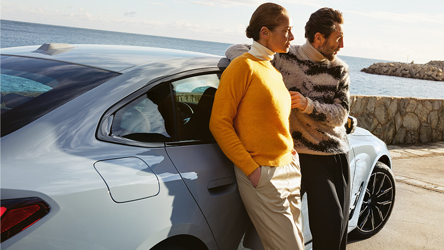 BMW Service Inclusive ilgums un līguma periodi jaunu automobiļu gadījumā BMW Service Inclusive, vīrietis un sieviete atbalstās pret BMW i4 uz piekrastes fona