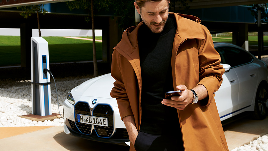 BMW Service Inclusive termiņš un līguma periodi elektroautomobiļu gadījumā BMW Service Inclusive, vīrietis skatās viedtālrunī uz BMW i4 fona uzlādes stacijā